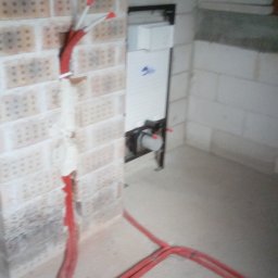 PRZEDSIĘBIORSTWO INSTALACYJNE SOTMET SPÓŁKA Z OGRANICZONĄ ODPOWIEDZIALNOŚCIĄ - Czerwone rury instalacji wodnej wystają ze ściany z cegły, prowadząc do podtynkowej toalety w surowym wnętrzu. Pianka montażowa uszczelnia przejścia rur.