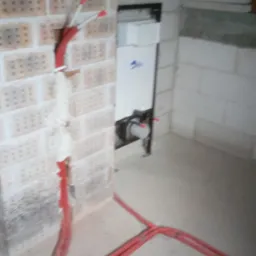 Czerwone rury instalacji wodnej wychodzące ze ściany z cegły i podłogi, w tle biała zabudowa podtynkowa WC w surowym pomieszczeniu.