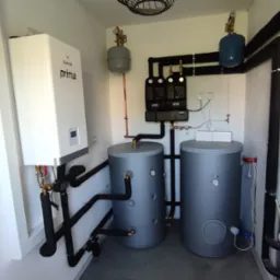 Kotłownia z białym piecem gazowym Galmet Prima, dwoma szarymi zbiornikami i czarnymi rurami w izolacji. Elementy instalacji zamontowane w pomieszczeniu z białymi ścianami.