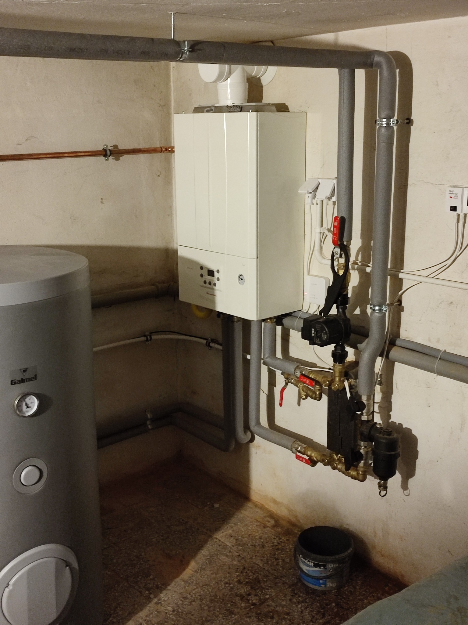 Biały piec gazowy zamontowany na ścianie w piwnicy, z widocznymi rurami, zaworami i szarym zasobnikiem ciepłej wody Galmet z boku.