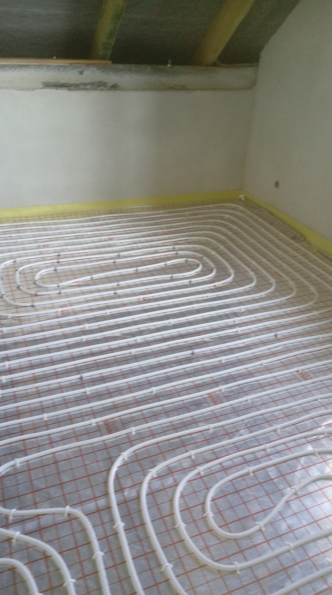 Białe rury ogrzewania podłogowego ułożone na folii z czerwoną siatką w jasnym pomieszczeniu, zabezpieczone żółtą taśmą przy ścianach. Widok z góry na instalację.