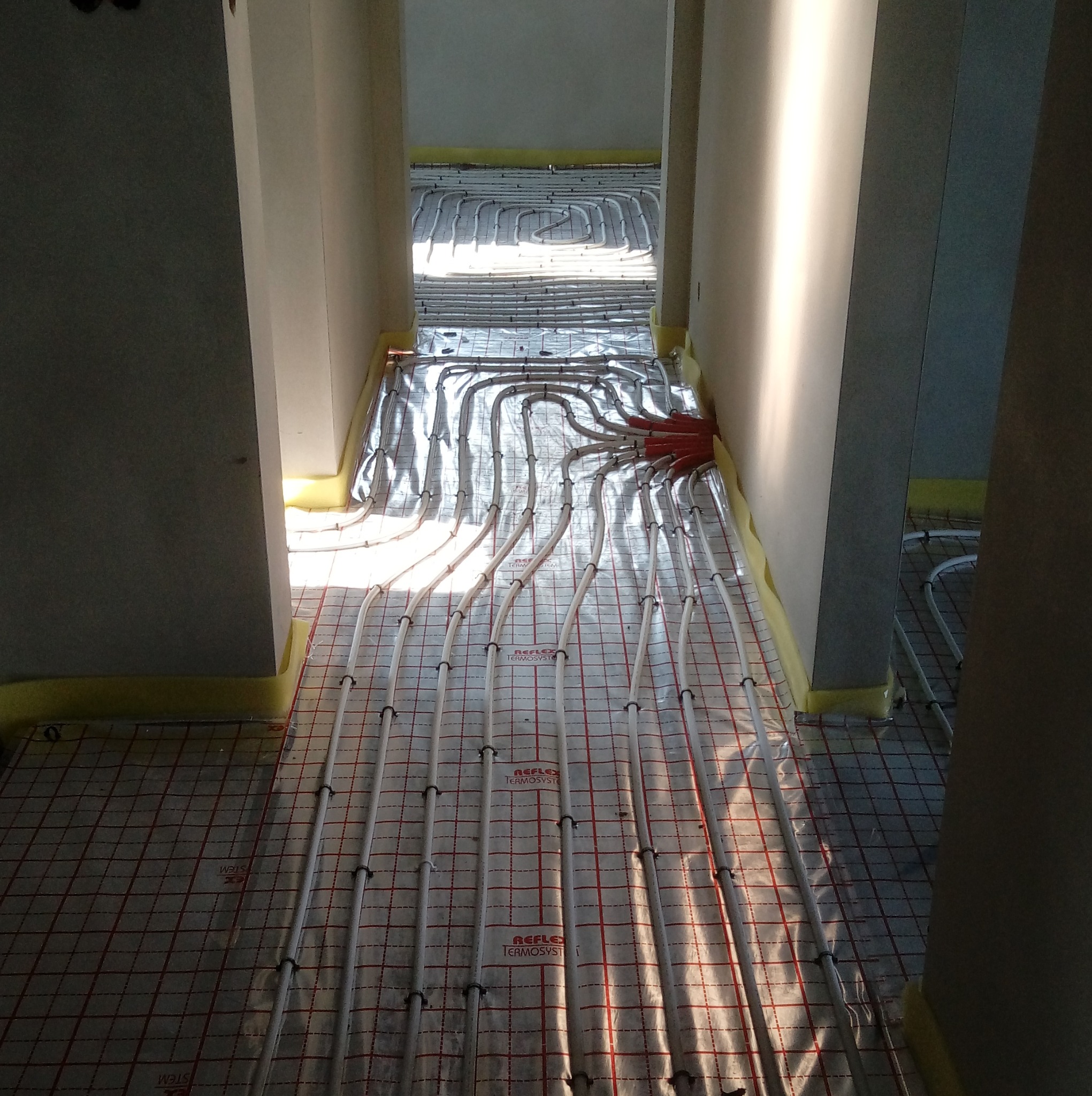 Białe rury ogrzewania podłogowego ułożone na folii z siatką, widoczne między ścianami korytarza w jasnym świetle. Etap instalacji przed wylewką.