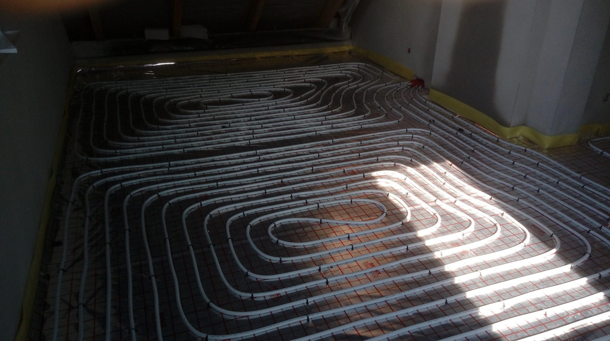 Białe rury ogrzewania podłogowego ułożone spiralnie na czerwonej siatce w jasnym pomieszczeniu, widok z góry, żółta folia przy ścianach.