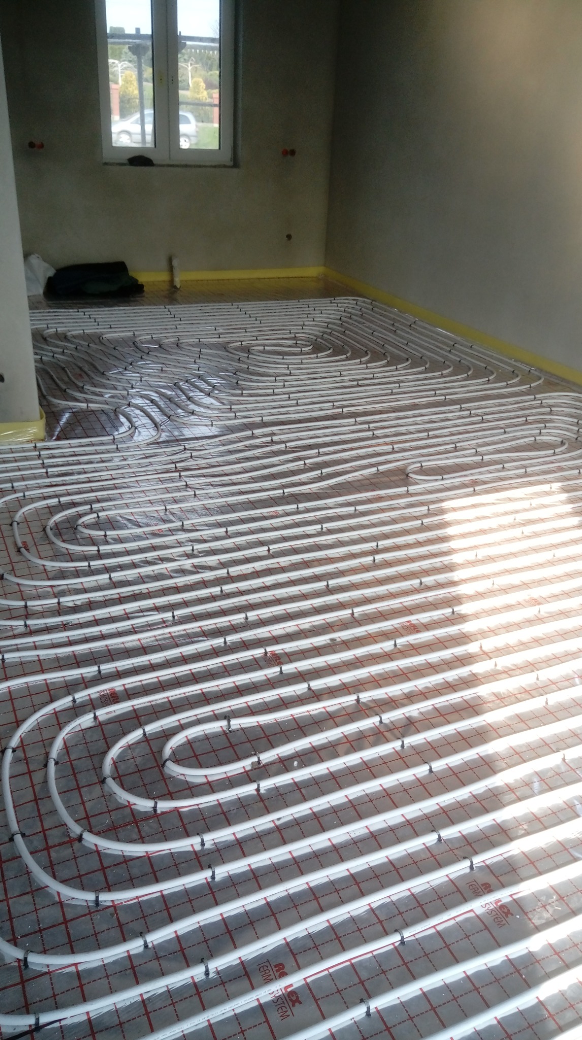 Ułożone rury ogrzewania podłogowego na folii z siatką w jasnym pomieszczeniu z oknem. Widoczne mocowania rur i żółta taśma izolacyjna przy ścianach.