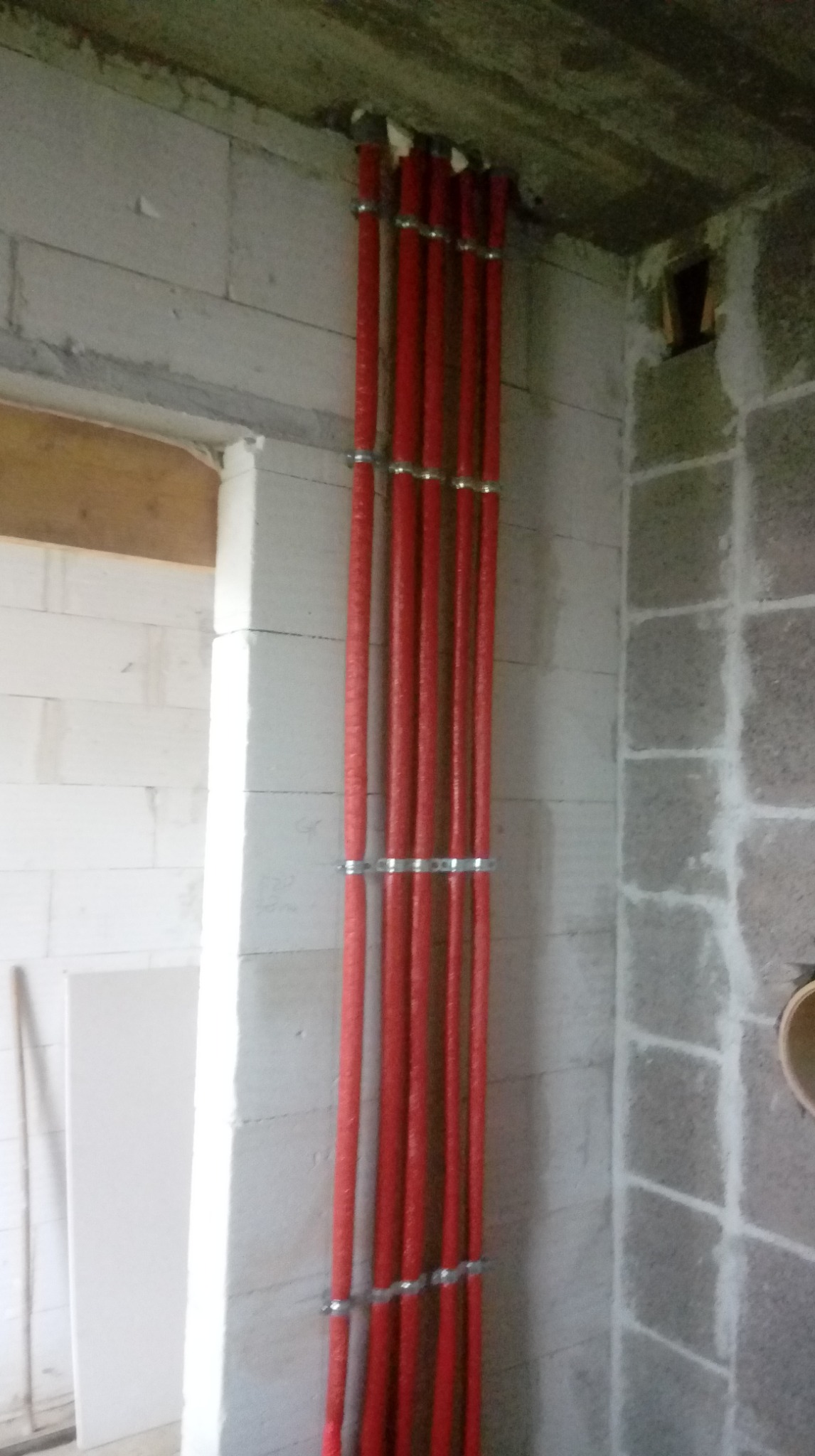 Czerwone, zaizolowane rury instalacji grzewczej, pionowo zamocowane do ściany z betonu komórkowego za pomocą metalowych uchwytów. Surowy stan wykończenia wnętrza.