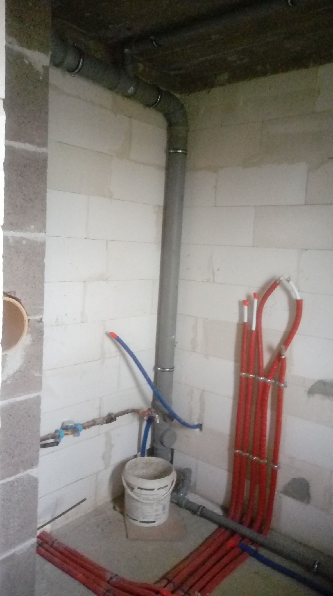 Instalacja wodno-kanalizacyjna w surowym wnętrzu: szare rury pionowe i poziome, czerwone rury ogrzewania podłogowego oraz białe ściany z betonu komórkowego.