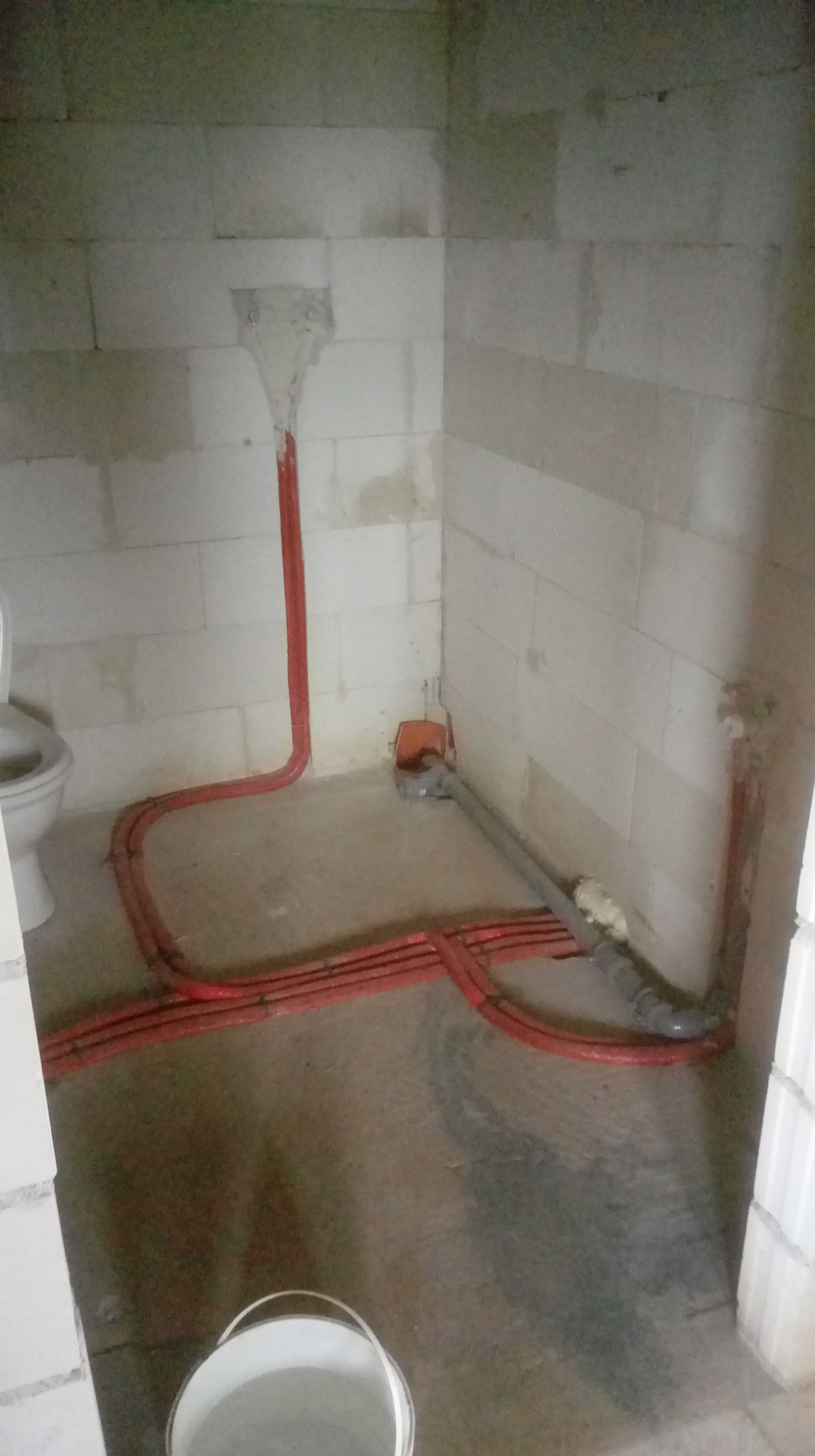 Surowy stan łazienki z widocznymi czerwonymi rurami instalacji wodnej i szarą rurą kanalizacyjną. Częściowo widoczna toaleta i biały kubeł na podłodze.