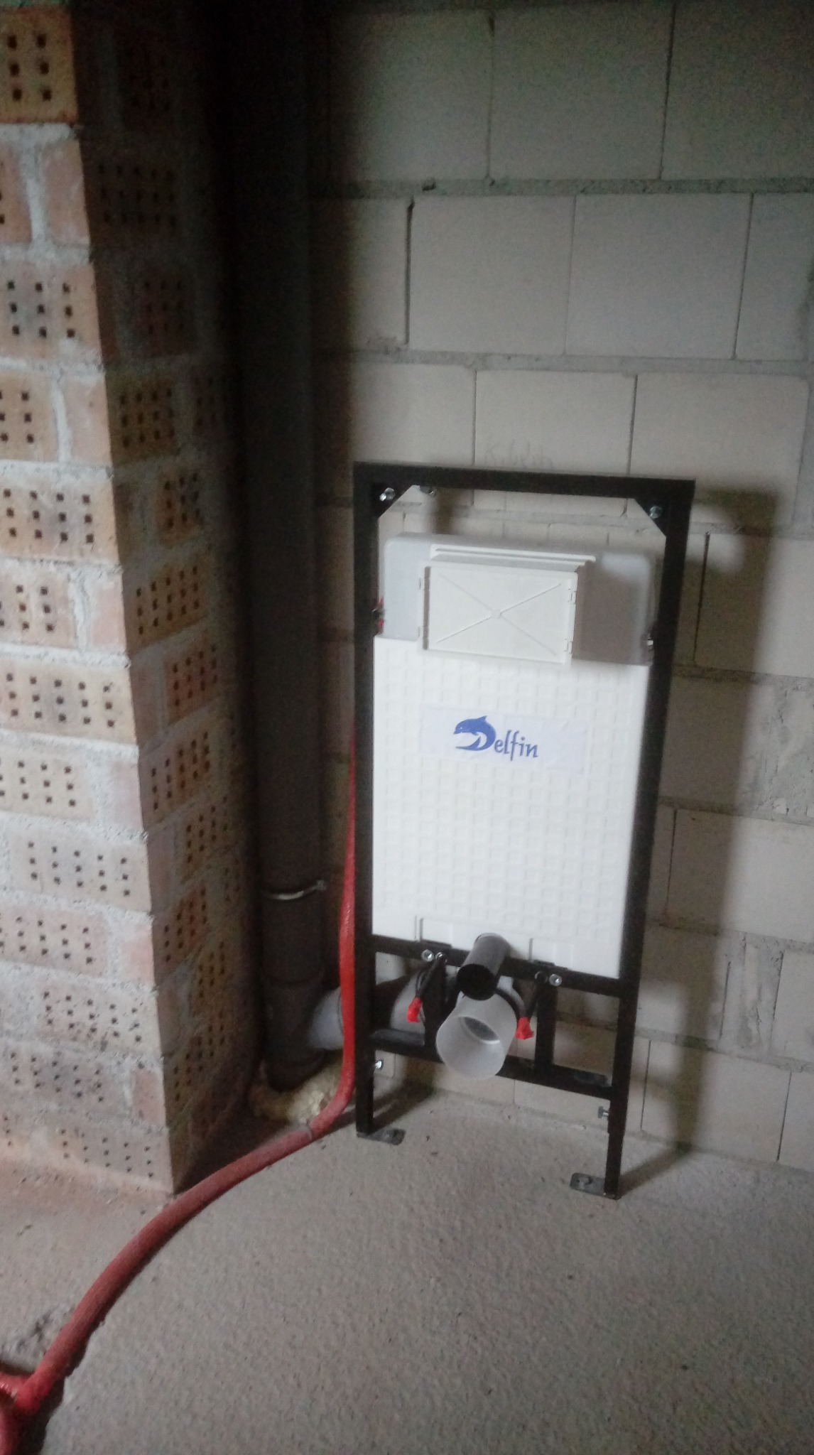Stelaż podtynkowy WC marki Delfin zamontowany przy ścianie z bloczków, z widocznymi rurami doprowadzającymi wodę i odpływowymi. Surowy stan wykończenia.