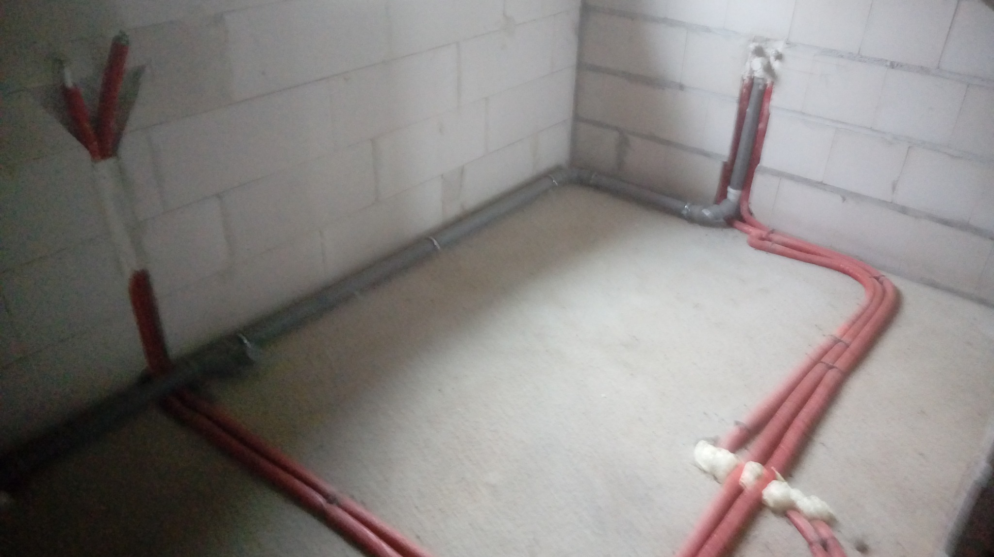 Szare i czerwone rury instalacyjne prowadzone wzdłuż ścian i podłogi pomieszczenia w stanie surowym. Widoczne łączenia i pianka montażowa.