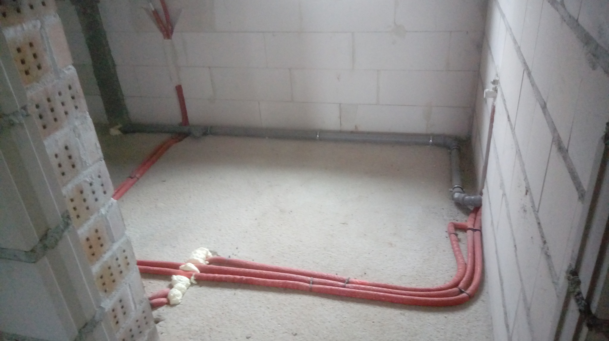 Wnętrze w trakcie prac instalacyjnych: czerwone rury biegną po podłodze wzdłuż ściany z betonu komórkowego, szara rura pod stropem. Widoczne pianka montażowa.