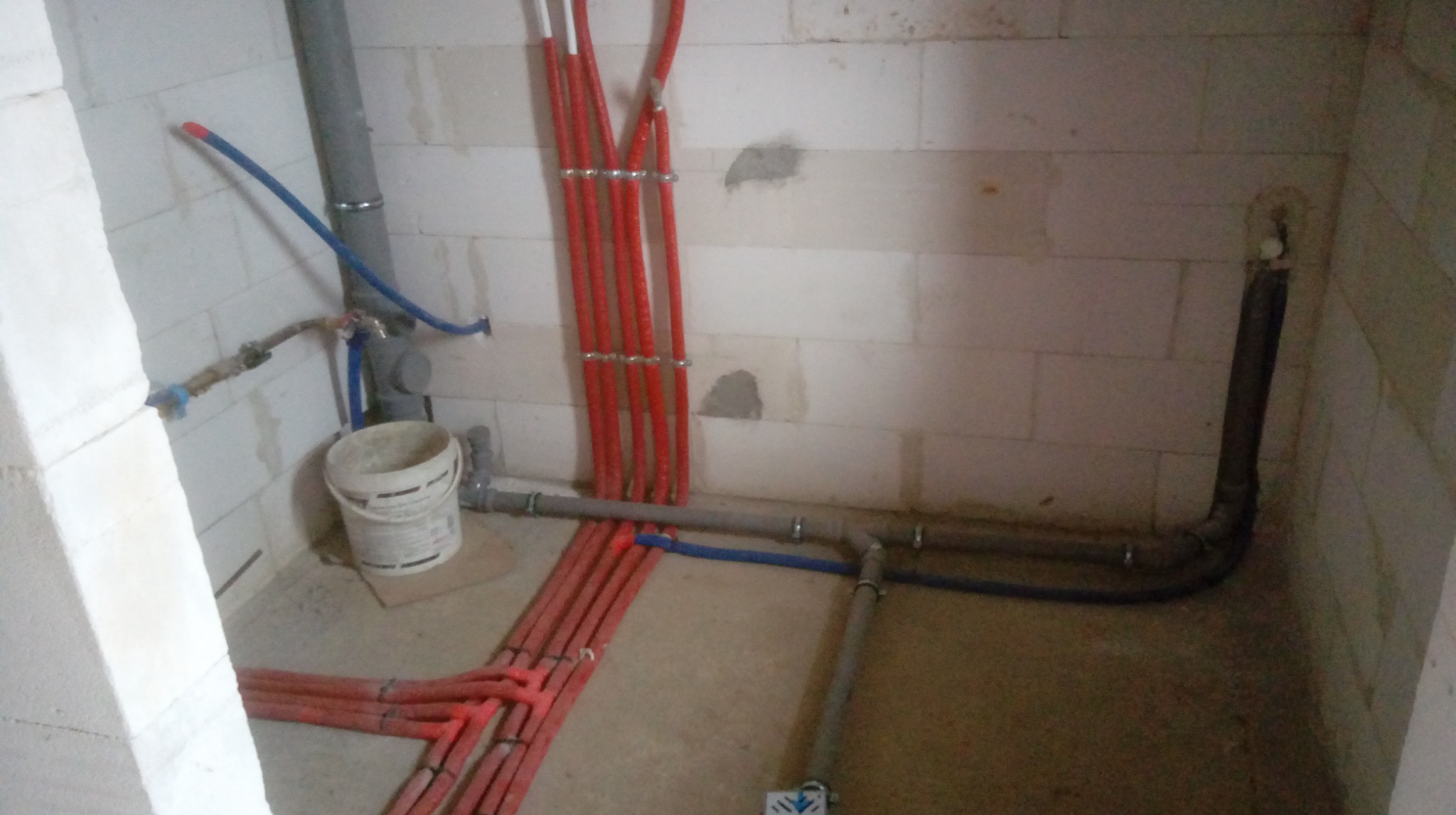 Instalacja wodna i kanalizacyjna w surowym stanie, z czerwonymi rurami ogrzewania podłogowego i szarymi rurami kanalizacyjnymi. Ściany z białych pustaków.