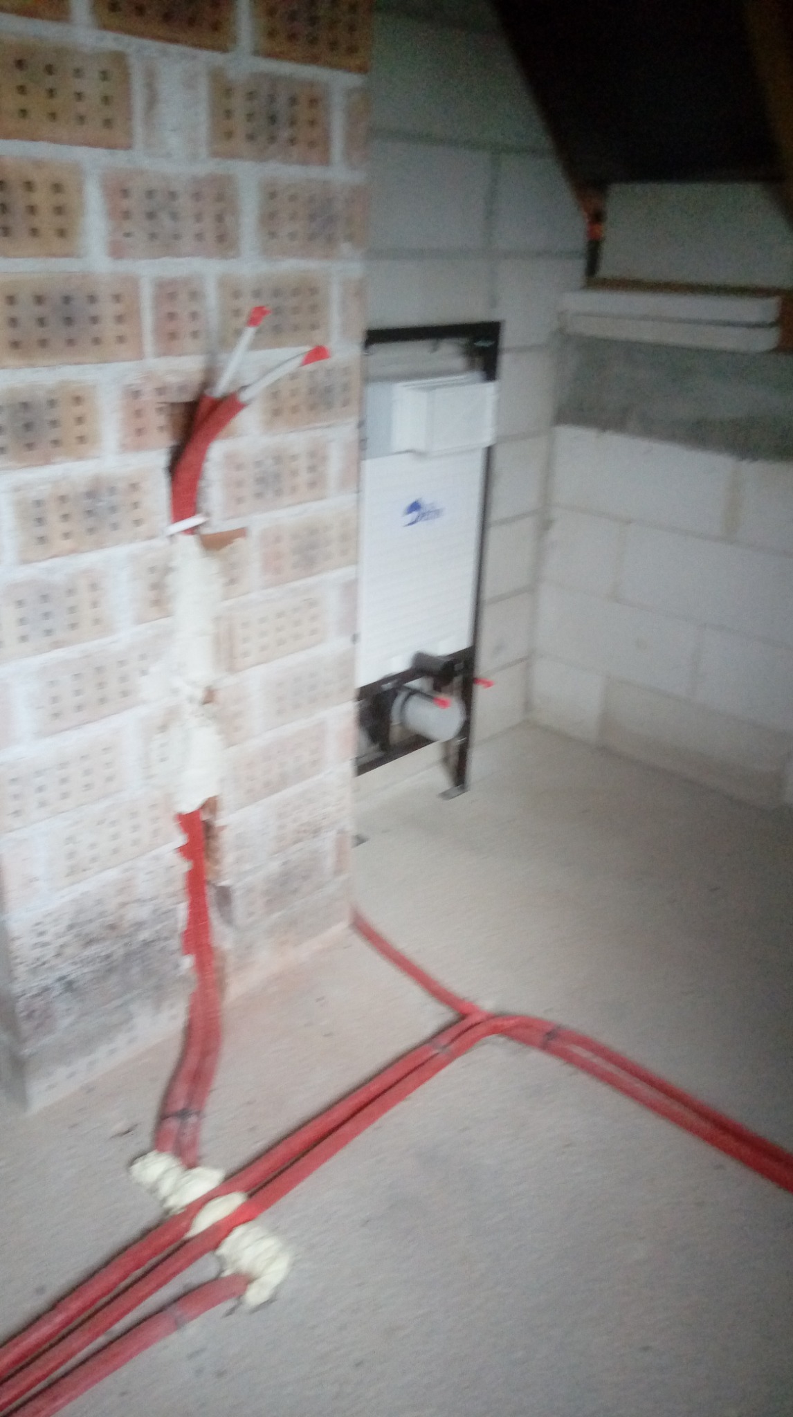 Czerwone rury instalacji wodnej wychodzące ze ściany z cegły i podłogi, w tle biała zabudowa podtynkowa WC w surowym pomieszczeniu.