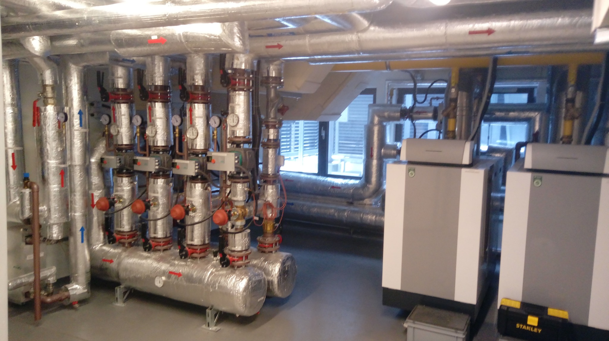 Złożona instalacja grzewcza z zaizolowanymi rurami i zaworami, manometry, dwa kotły grzewcze z żółtym przewodem gazowym, skrzynka narzędziowa Stanley.