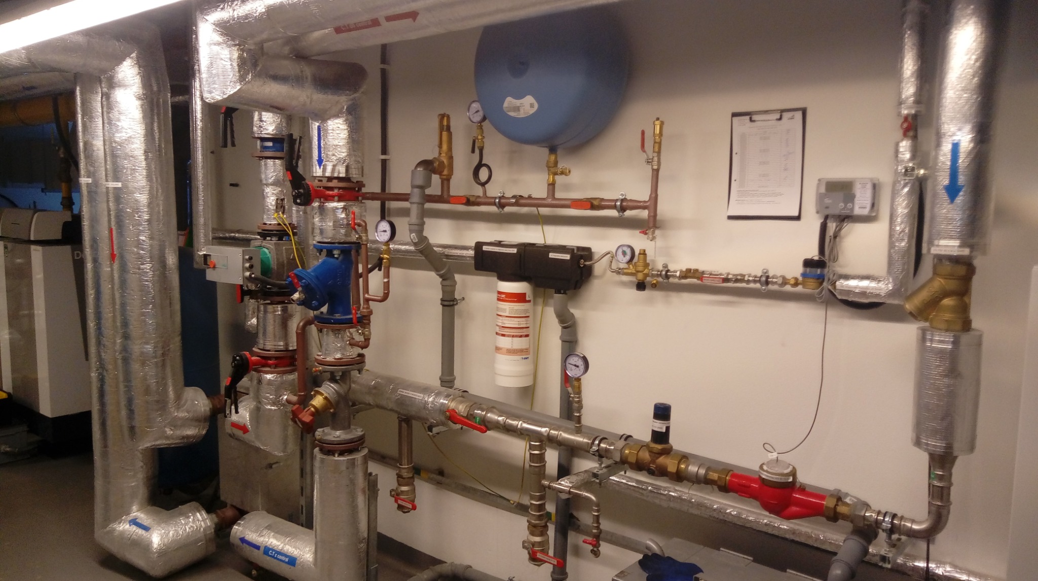 Złożona instalacja grzewcza z licznymi rurami, zaworami, manometrami i filtrem, starannie zamontowana na ścianie w pomieszczeniu technicznym. Rury zaizolowane.