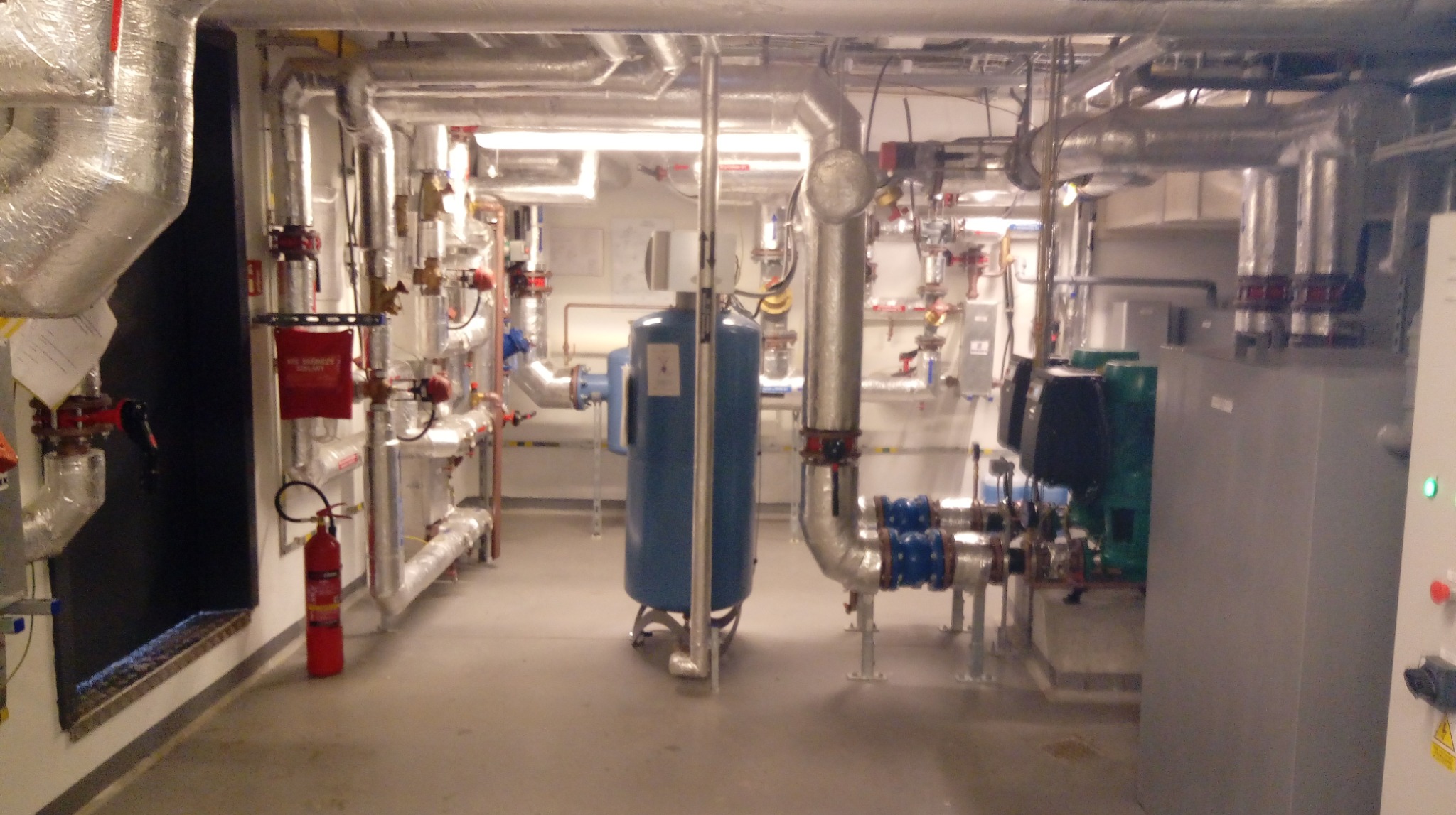 Węzeł cieplny z licznymi rurami, zaworami i zbiornikami. Widoczny zbiornik wyrównawczy, pompy obiegowe oraz instalacja przeciwpożarowa z gaśnicą.