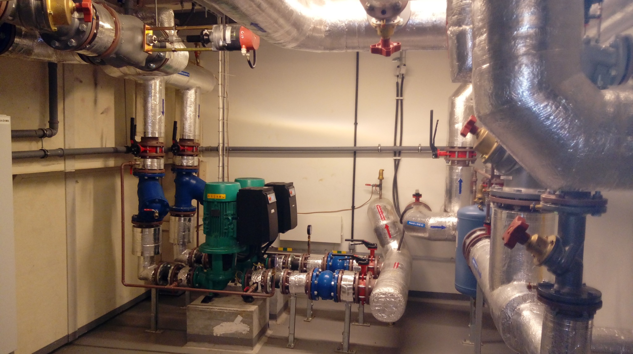 Węzeł cieplny z zielonymi pompami i licznymi rurami, zaworami oraz elementami izolacji termicznej. Widoczne różne kolory rur i zaworów.