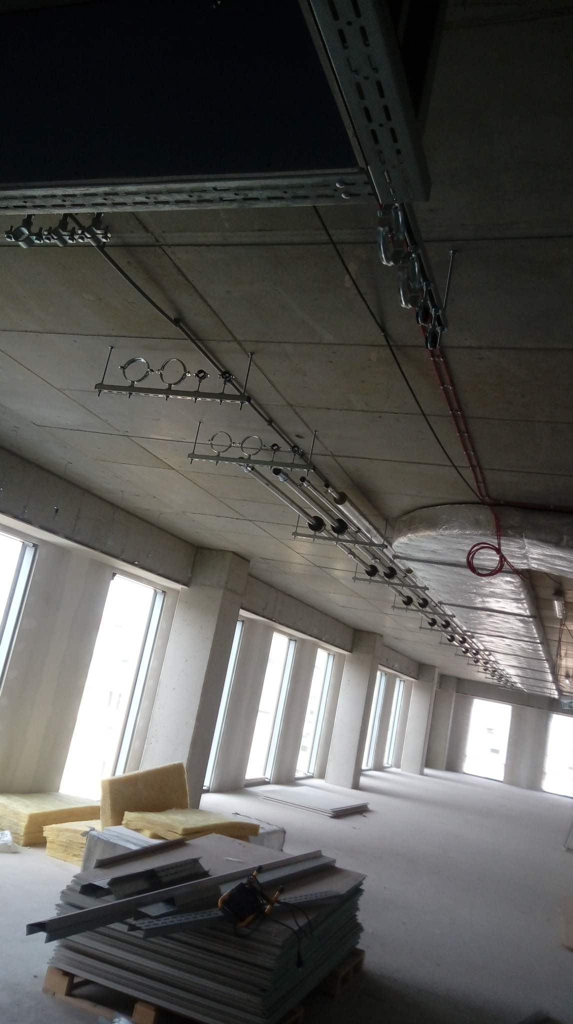 Wnętrze w trakcie instalacji: widoczne rury, kable i przewody pod sufitem, pod oknami materiały izolacyjne i płyty. Surowy stan, duże okna, wysokie pomieszczenie.
