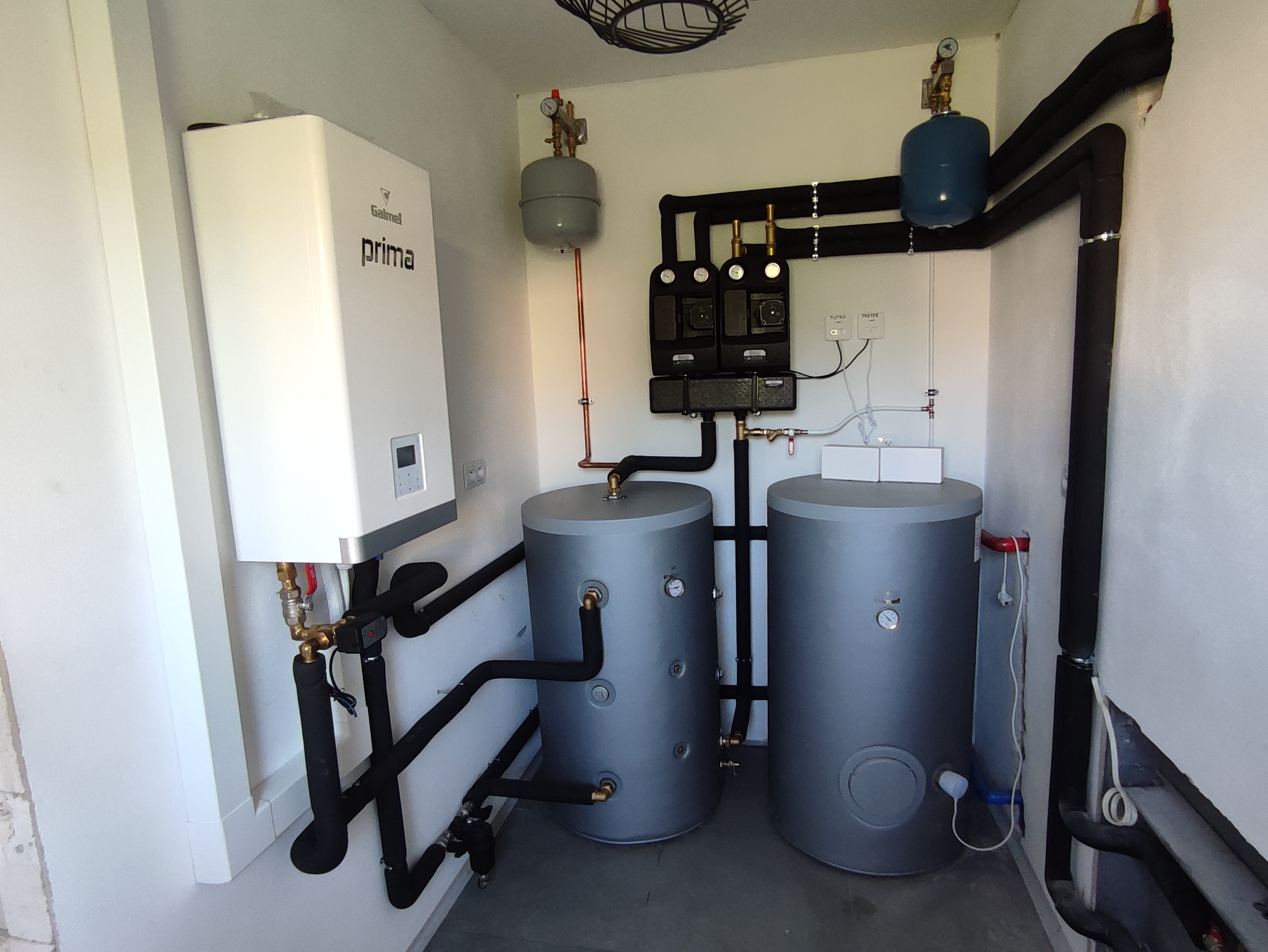 Kotłownia z białym piecem gazowym Galmet Prima, dwoma szarymi zbiornikami i czarnymi rurami w izolacji. Elementy instalacji zamontowane w pomieszczeniu z białymi ścianami.
