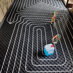 LULU Volha Vishniakova - Białe rurki ogrzewania podłogowego ułożone na czarnej macie z wypustkami, widok z góry. W tle surowe ściany w trakcie remontu. Kanister z płynem i narzędzia obok.