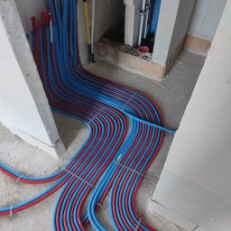 LULU Volha Vishniakova - Równolegle ułożone, czerwono-niebieskie rury instalacyjne podłogowe, przymocowane metalowymi klamrami, na tle surowego betonu i bloczków. Widoczne fragmenty pionowych instalacji.