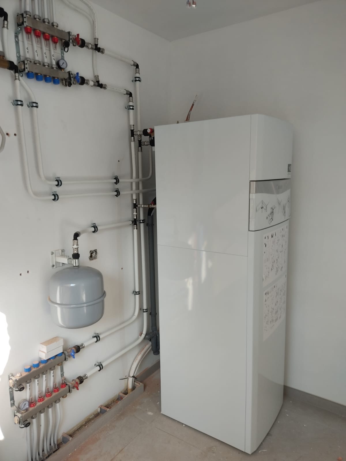 Biały piec grzewczy z podłączonymi rurami instalacji centralnego ogrzewania, widoczne elementy regulacyjne i zbiornik wyrównawczy. Białe ściany, surowa podłoga.