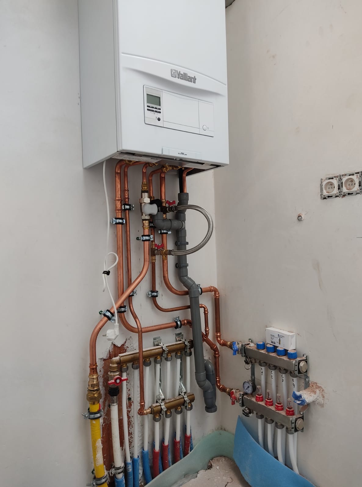 Biały piec Vaillant z miedzianymi rurami i szarymi elementami instalacji, zamontowany na ścianie w surowym stanie. Widoczne rozdzielacze i podłączenia.