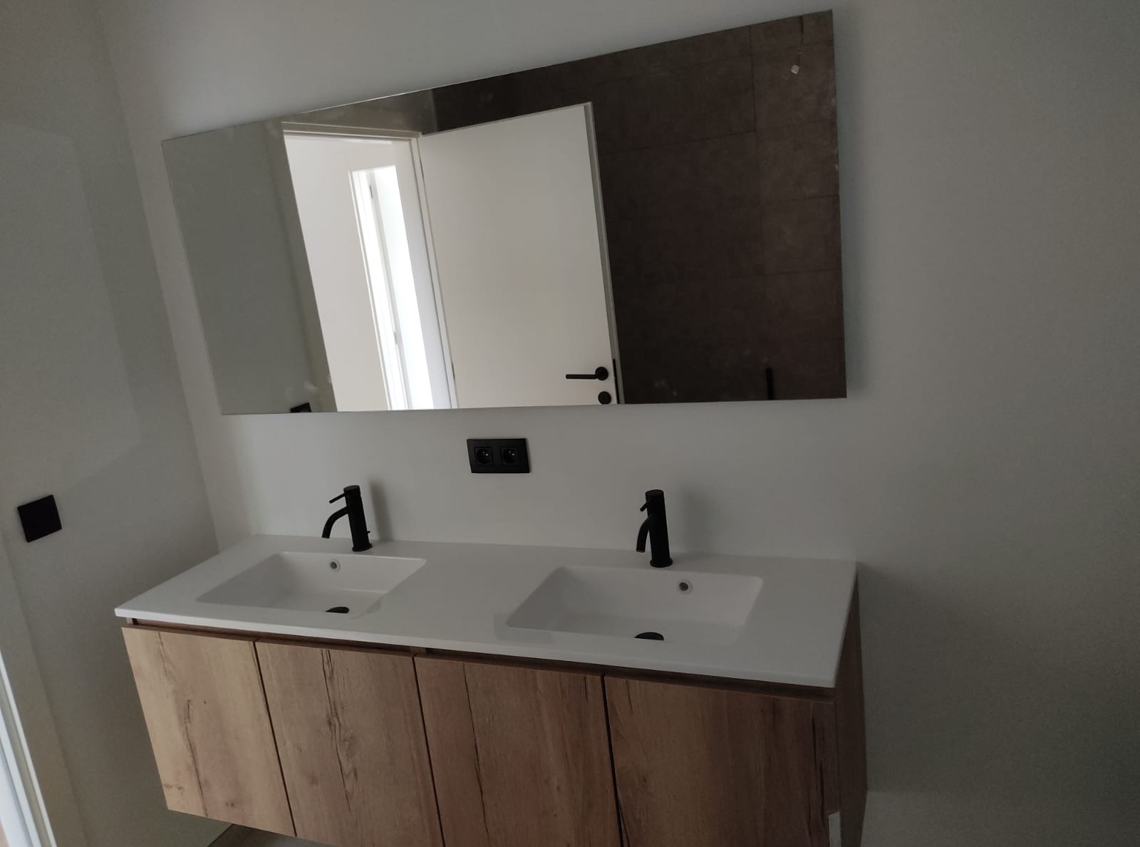 Nowoczesna łazienka z podwójną umywalką nablatową, czarne baterie, lustro z widokiem na drzwi i fragment ściany z brązowymi płytkami. Drewnopodobna szafka pod umywalką.