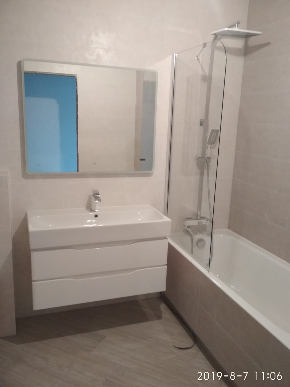 Nowoczesna łazienka z wiszącą szafką umywalkową, lustrem z podświetleniem LED i kabiną prysznicową nad wanną, wykończona płytkami w odcieniach beżu.