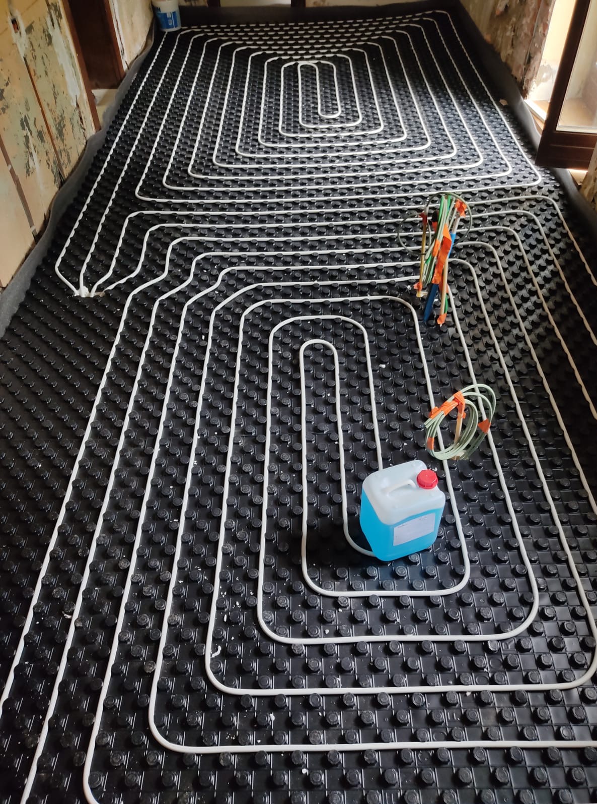 Białe rurki ogrzewania podłogowego ułożone na czarnej macie z wypustkami, widok z góry. W tle surowe ściany w trakcie remontu. Kanister z płynem i narzędzia obok.