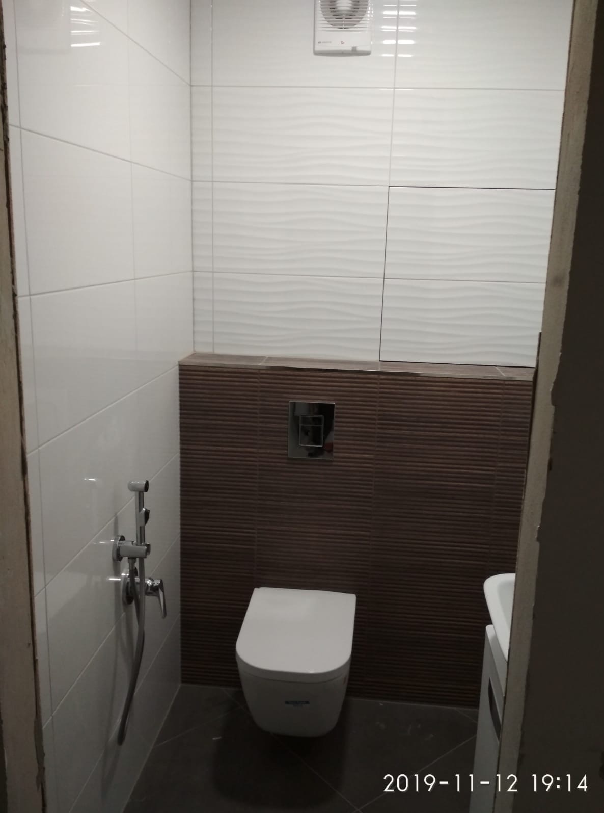 Nowoczesna toaleta po remoncie z białymi i brązowymi płytkami, widoczna armatura i wentylator. Ujęcie z narożnika pomieszczenia, z datą na dole zdjęcia.