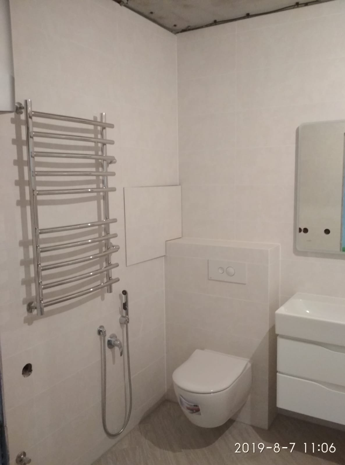 Wykończona łazienka z grzejnikiem drabinkowym, toaletą podwieszaną, umywalką z szafką i bidetką. Widoczne płytki ścienne i podłogowe w jasnych odcieniach.