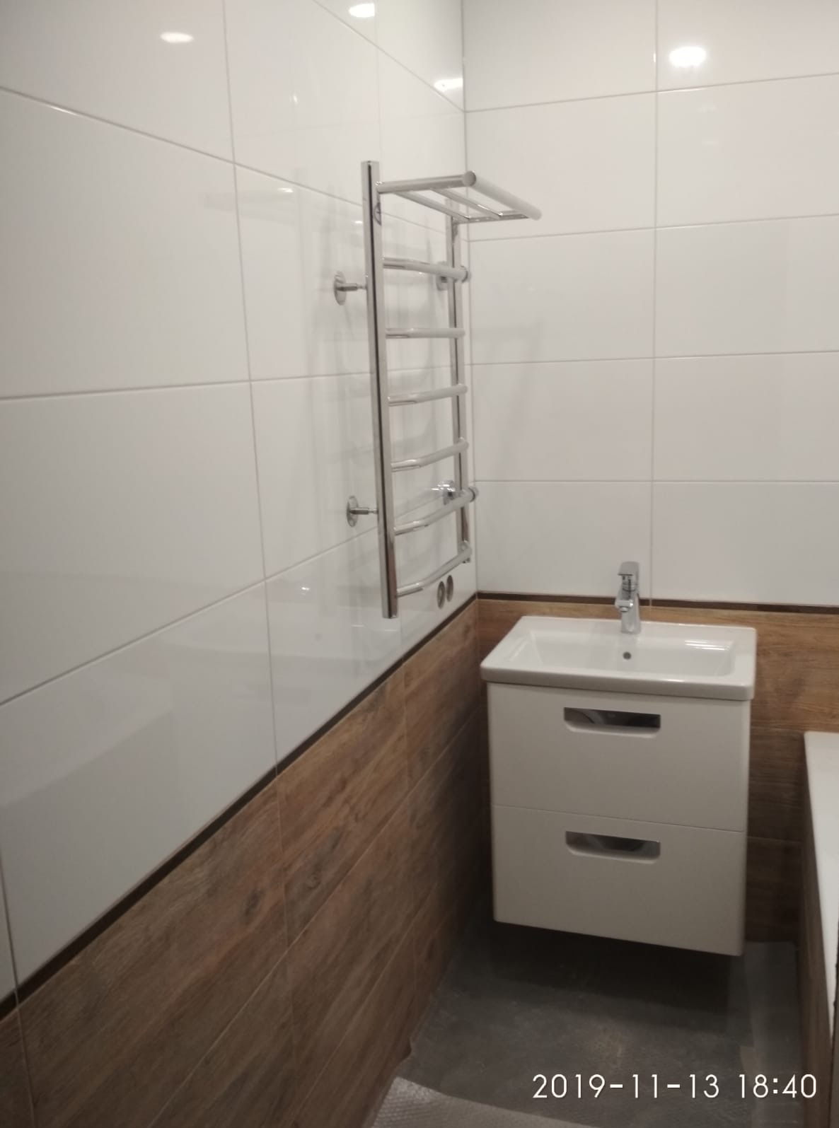 Wykończona łazienka: białe płytki, drewnopodobne panele, umywalka z szafką i chromowany grzejnik drabinkowy. Nowoczesny design, staranne wykonanie.