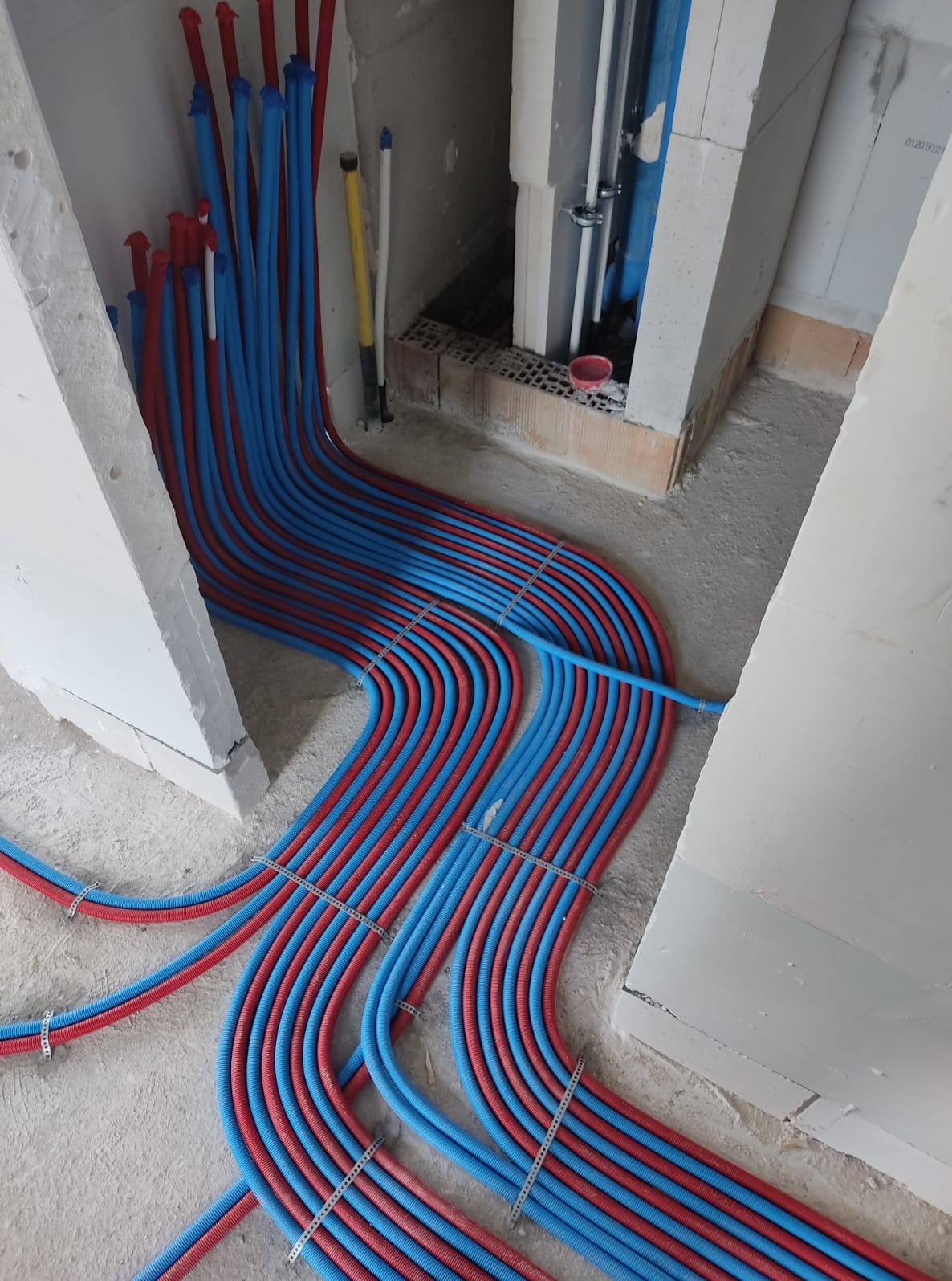 Równolegle ułożone, czerwono-niebieskie rury instalacyjne podłogowe, przymocowane metalowymi klamrami, na tle surowego betonu i bloczków. Widoczne fragmenty pionowych instalacji.