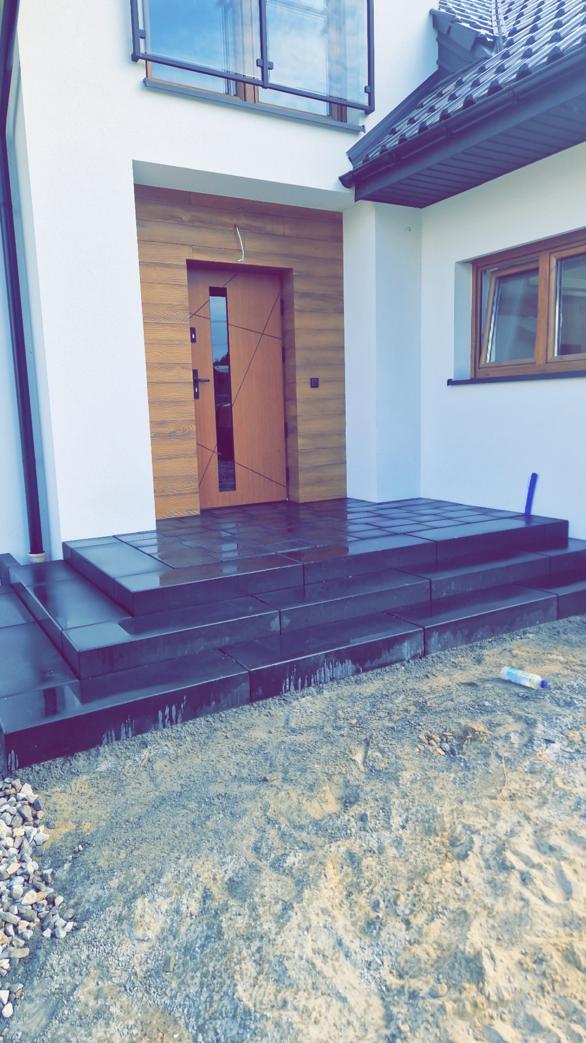 Nowoczesne, ciemne schody z kostki brukowej prowadzące do wejścia domu z drewnianą elewacją. Widoczne ślady wilgoci i rozsypany żwir.