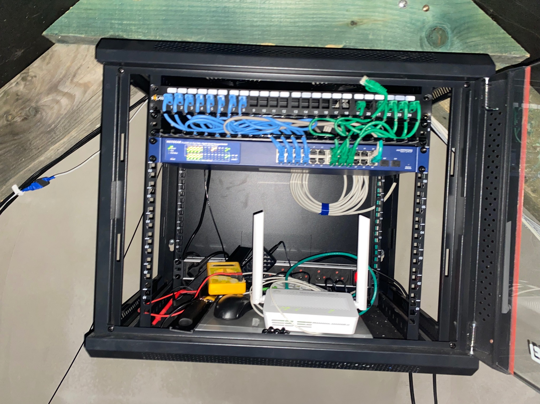 Otwarta szafa rack z patch panelem, routerem i okablowaniem w kolorach niebieskim i zielonym. Widoczny miernik i myszka komputerowa. Elementy sieci w domowym otoczeniu.