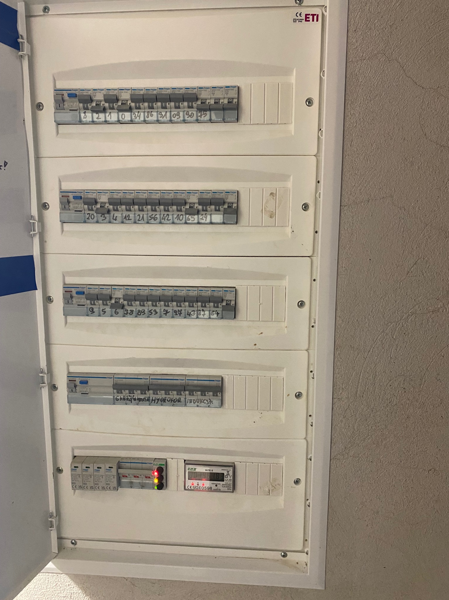 Rozdzielnia elektryczna z bezpiecznikami i licznikiem, widoczne oznaczenia i numery. Biała obudowa na szarej ścianie, detale instalacji.