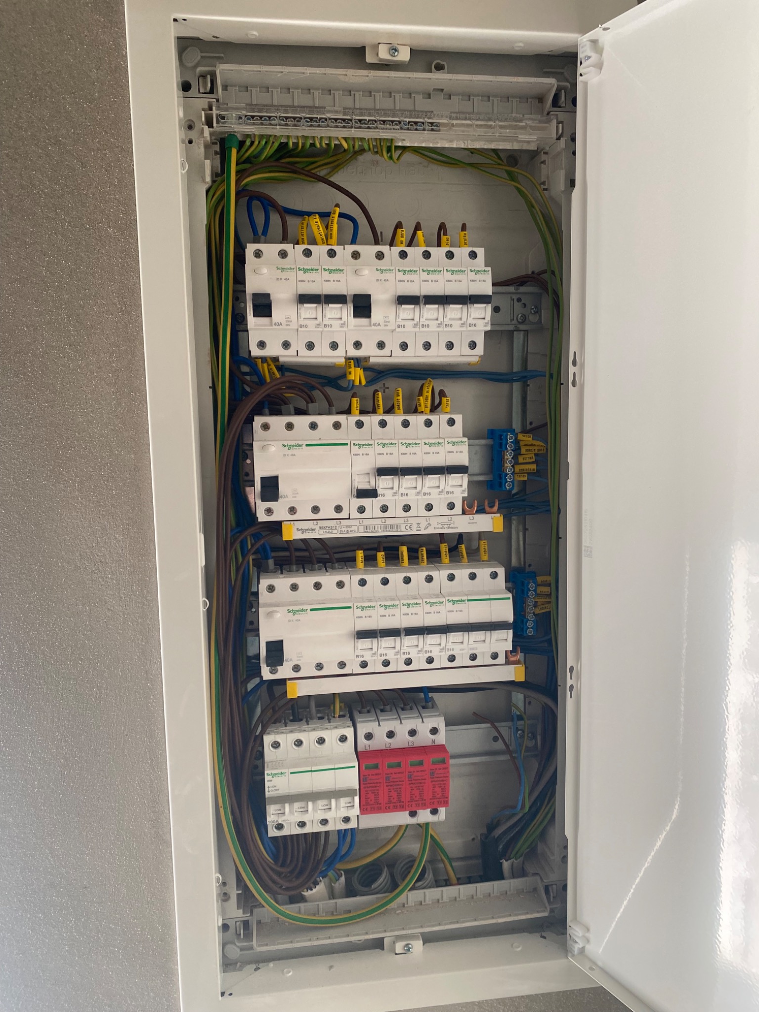 Otwarta, biała skrzynka rozdzielcza z bezpiecznikami i okablowaniem Schneider Electric. Widoczne brązowe, zielono-żółte i niebieskie przewody.