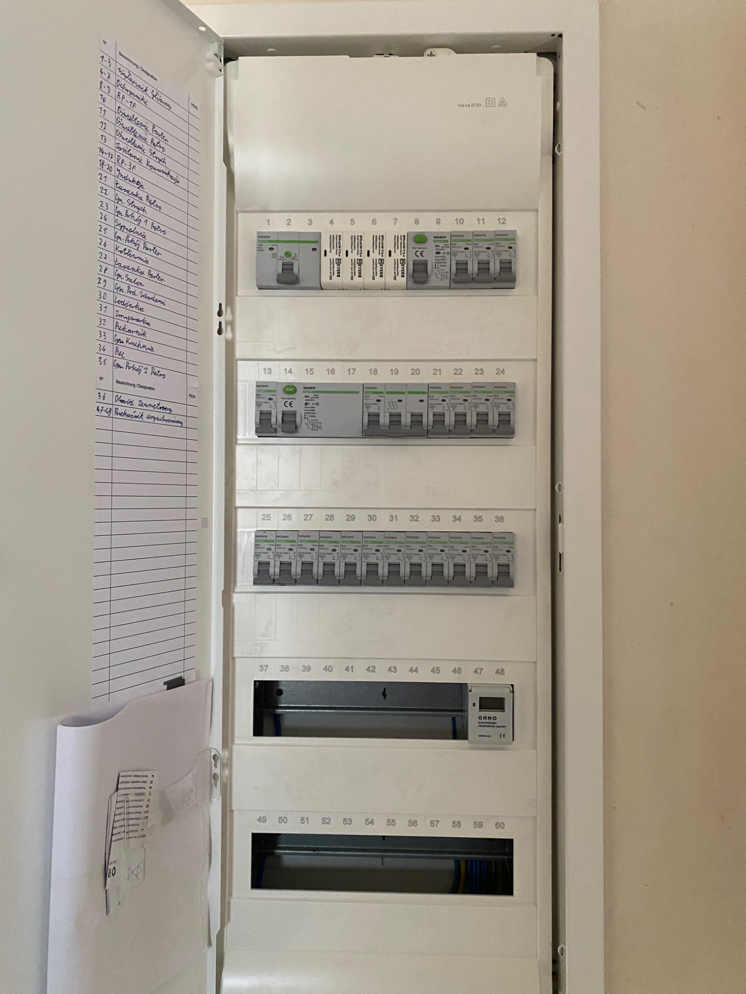 Otwarta skrzynka elektryczna z bezpiecznikami, widoczne opisy obwodów na kartce obok. Instalacja wewnątrz, numery bezpieczników od 1 do 60.