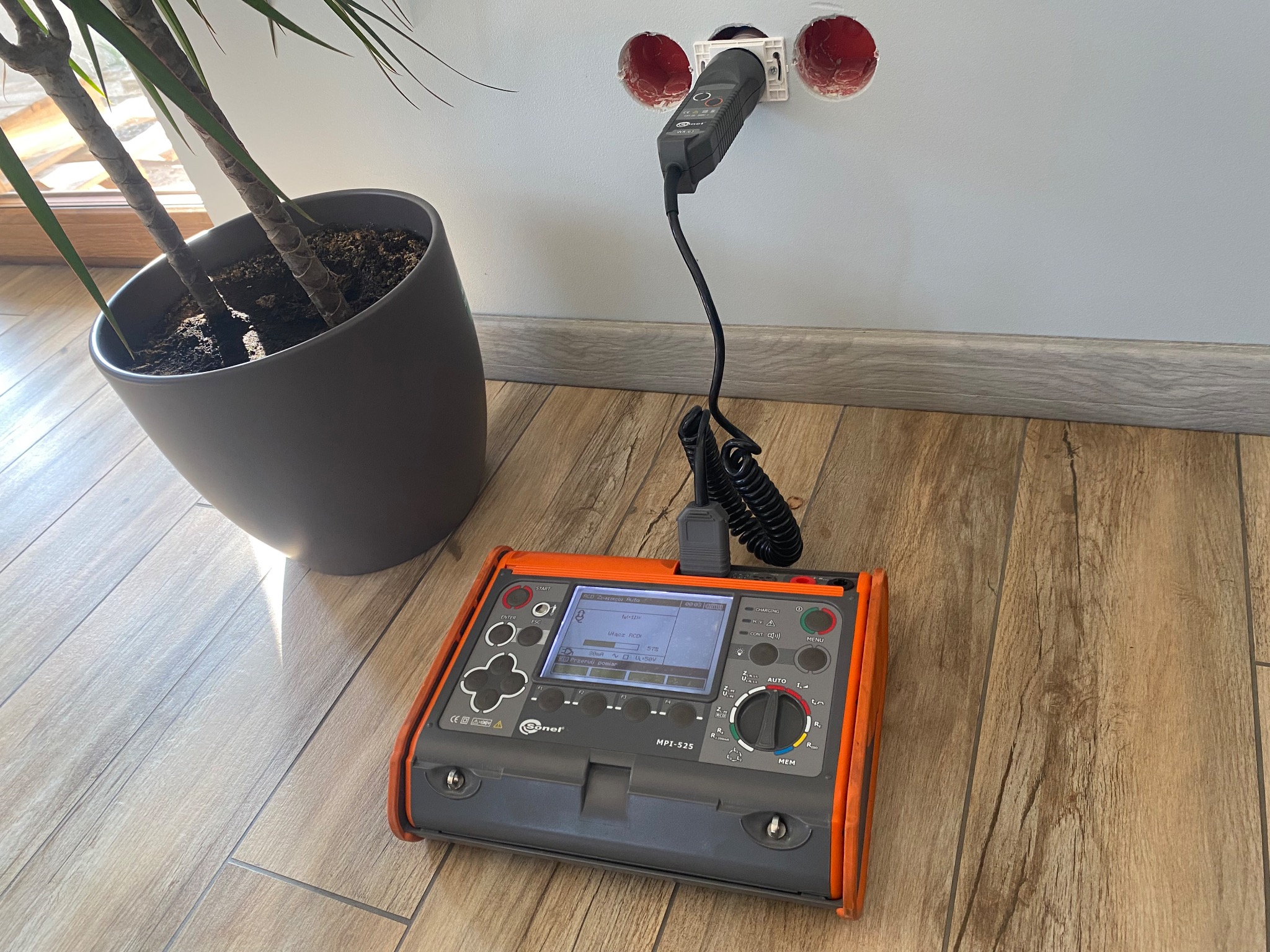 Pomiar gniazdka elektrycznego miernikiem Sonel MPI-525 na tle ściany z dwoma otworami pod gniazdka i rośliny doniczkowej. Widok z góry na podłodze z paneli.