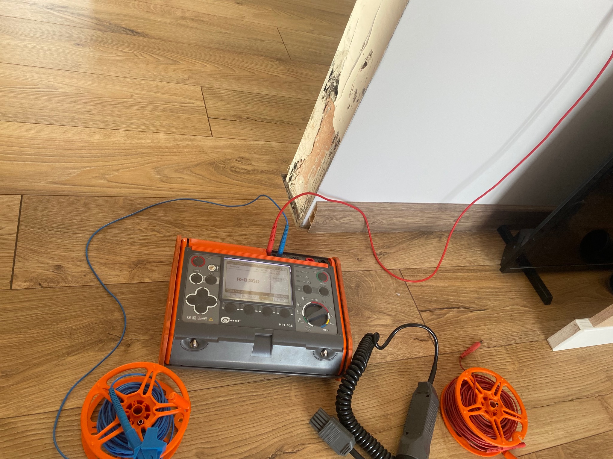 Miernik Sonel MPI-525 z podłączonymi przewodami pomiarowymi na drewnianej podłodze, obok szpule z przewodami. Pomiar rezystancji izolacji przy ścianie.
