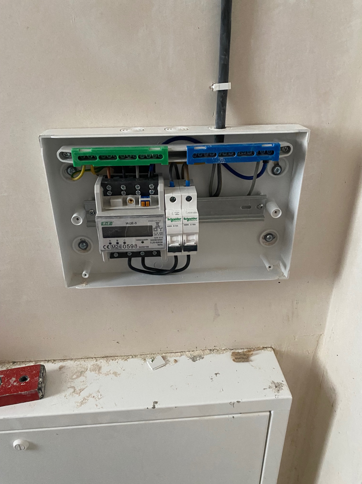 Biała skrzynka z licznikiem i bezpiecznikami Schneider Electric na ścianie. Widoczne przewody w kolorach zielonym i niebieskim. Elementy instalacji elektrycznej wewnątrz budynku.