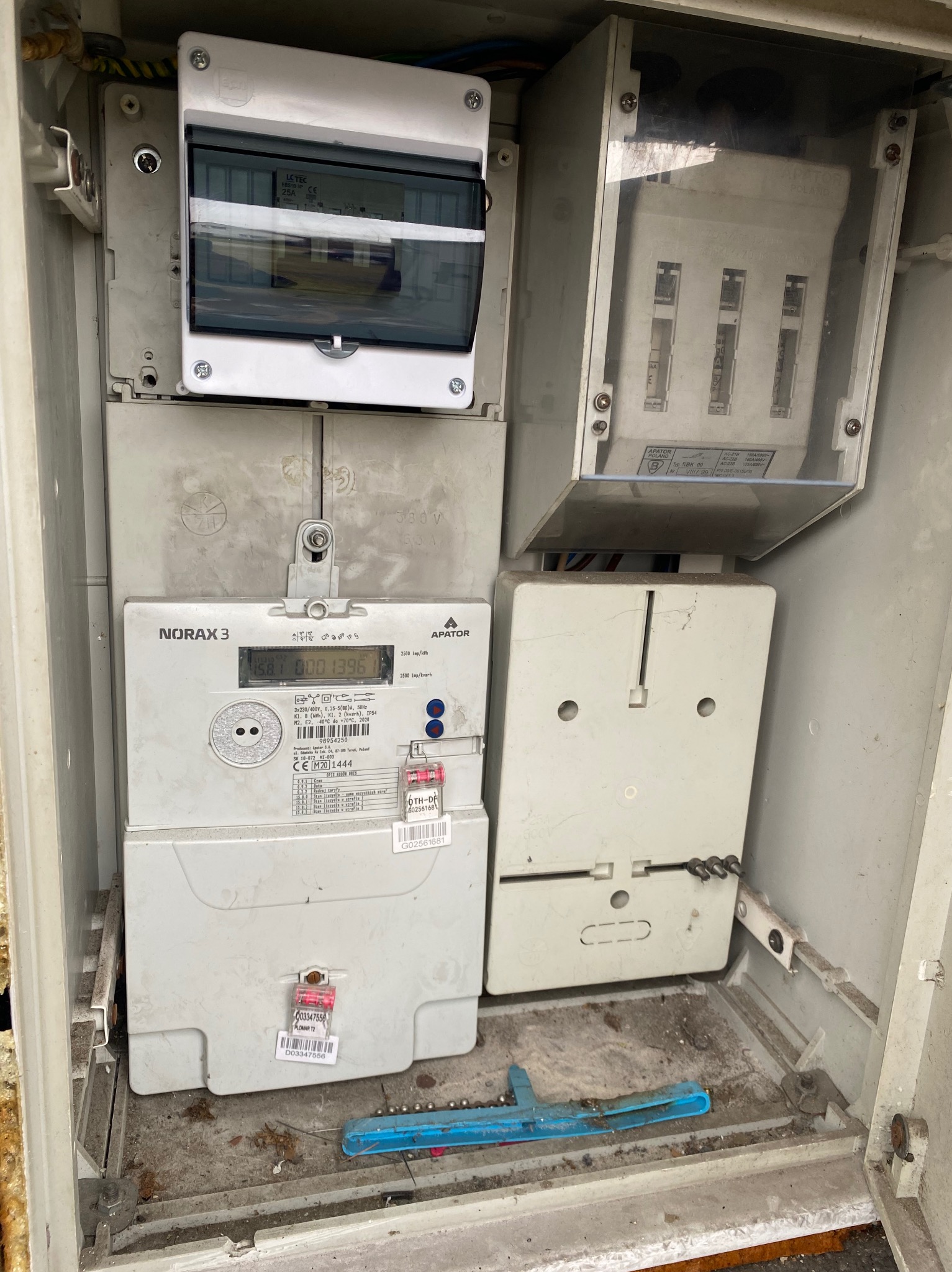 Skrzynka z licznikami energii elektrycznej i zabezpieczeniami, widoczne liczniki NORAX 3 i APATOR, plomby, przewody. Ujęcie z bliska, wewnątrz skrzynki.