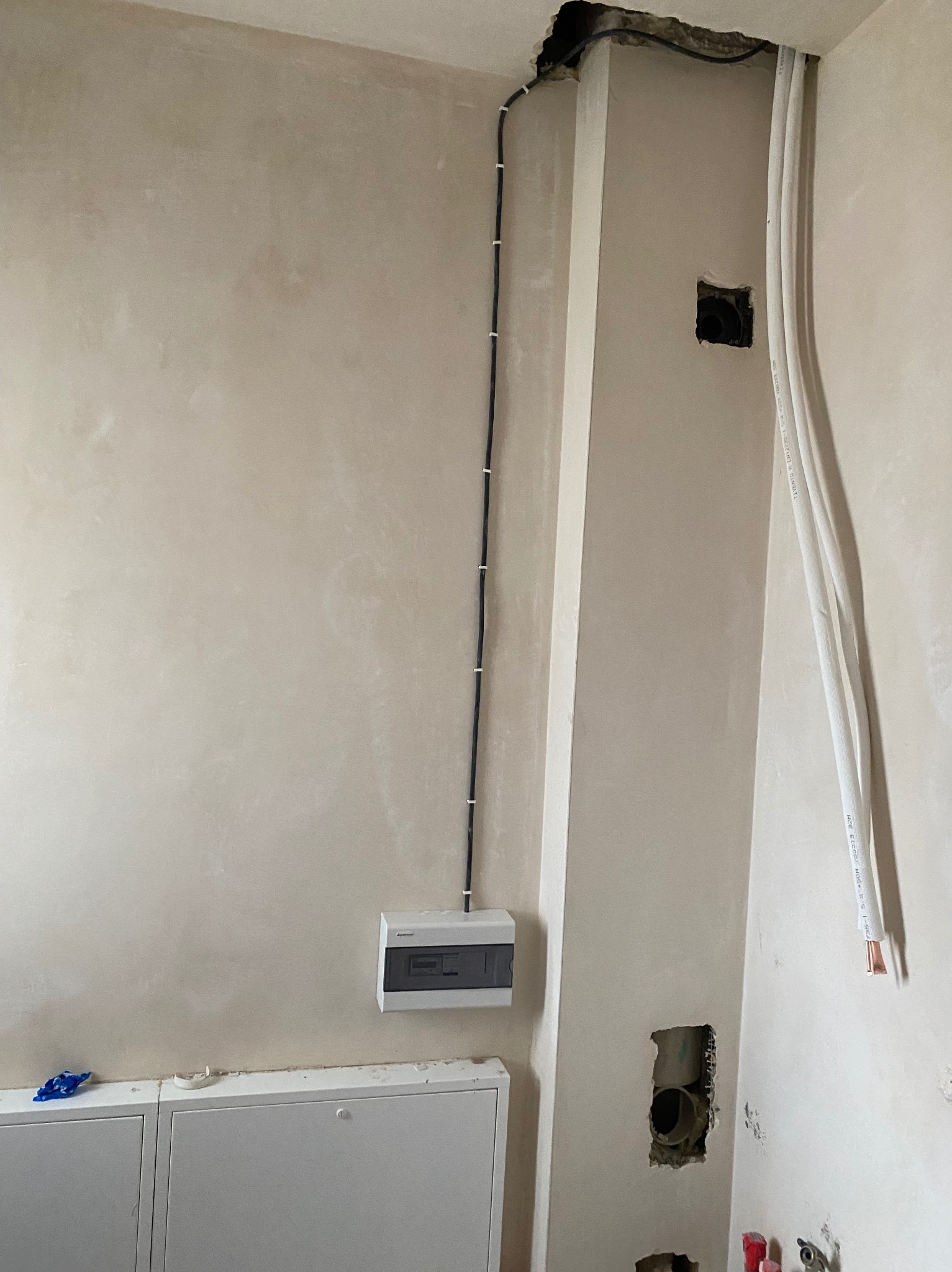 Szara ściana w trakcie remontu z widoczną puszką elektryczną, przewodami w listwach i otworami na instalacje. Widoczne ślady po kuciu i niedoskonałości tynku.