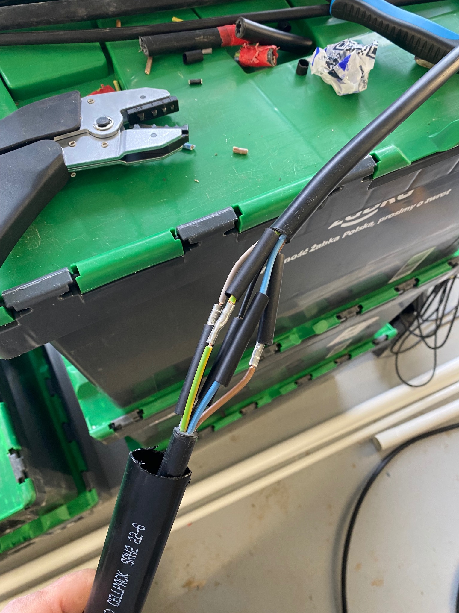Przygotowany kabel elektryczny z zaciśniętymi końcówkami, widoczne kolory przewodów. Obok leżą narzędzia do zaciskania. Tło stanowią zielone pojemniki.
