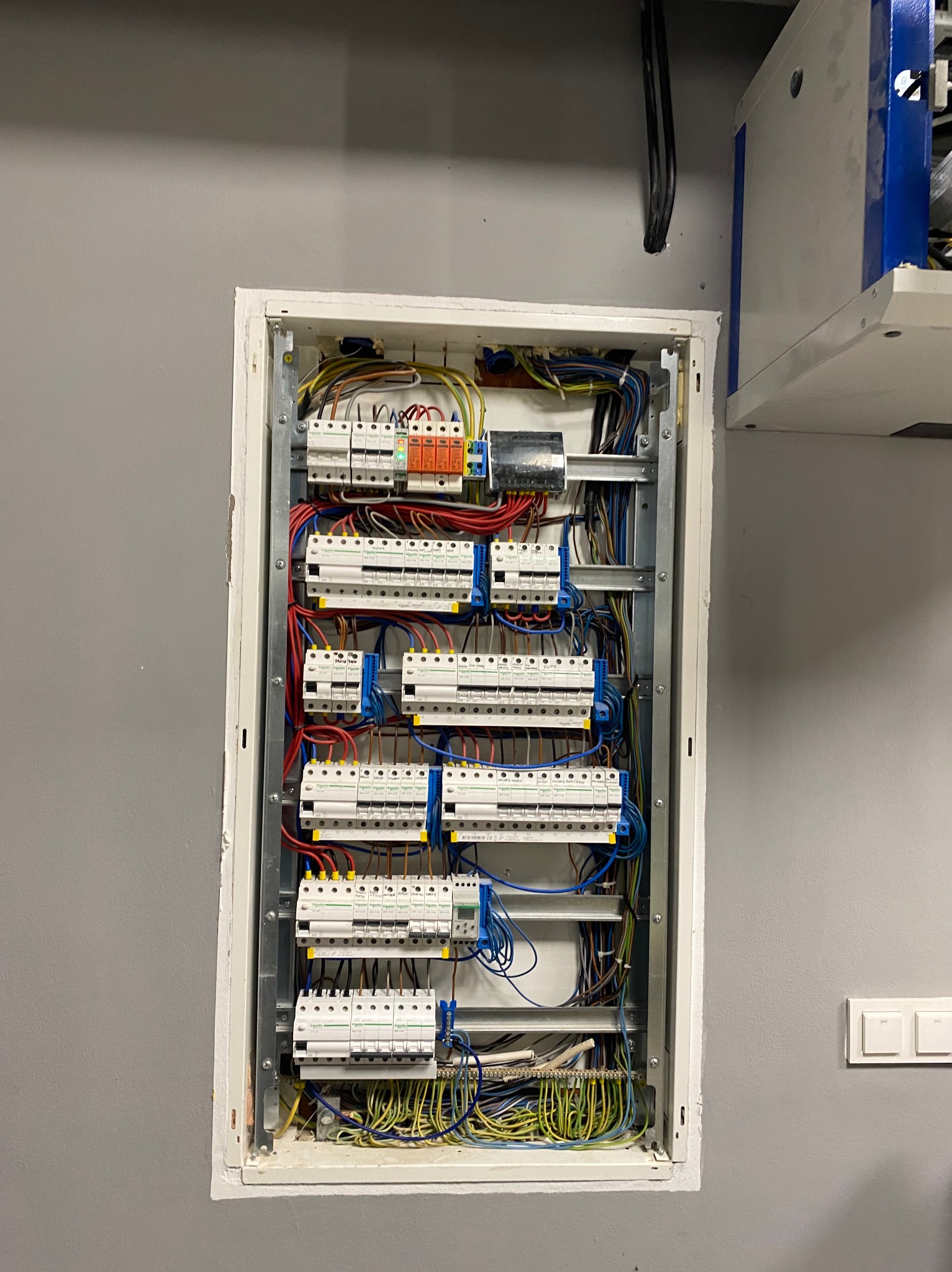 Rozdzielnica elektryczna w białej obudowie, zamontowana na szarej ścianie. Widoczne liczne przewody i zabezpieczenia. U dołu listwa zaciskowa z przewodami.