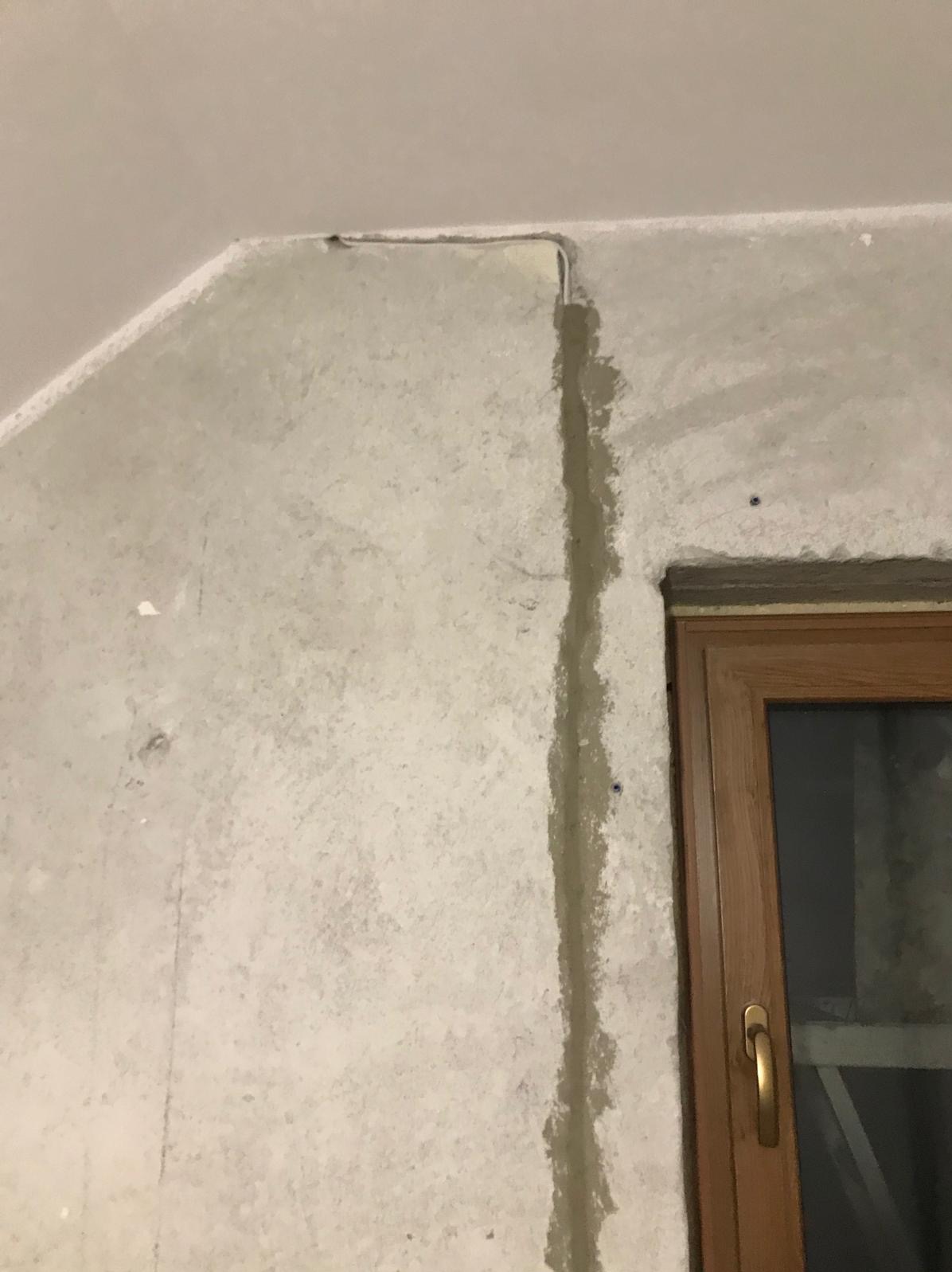 Szara ściana z bruzdą wypełnioną gipsem, w której poprowadzono przewód. Widoczne okno z drewnianą ramą. Ujęcie z bliska, surowy stan wykończenia.