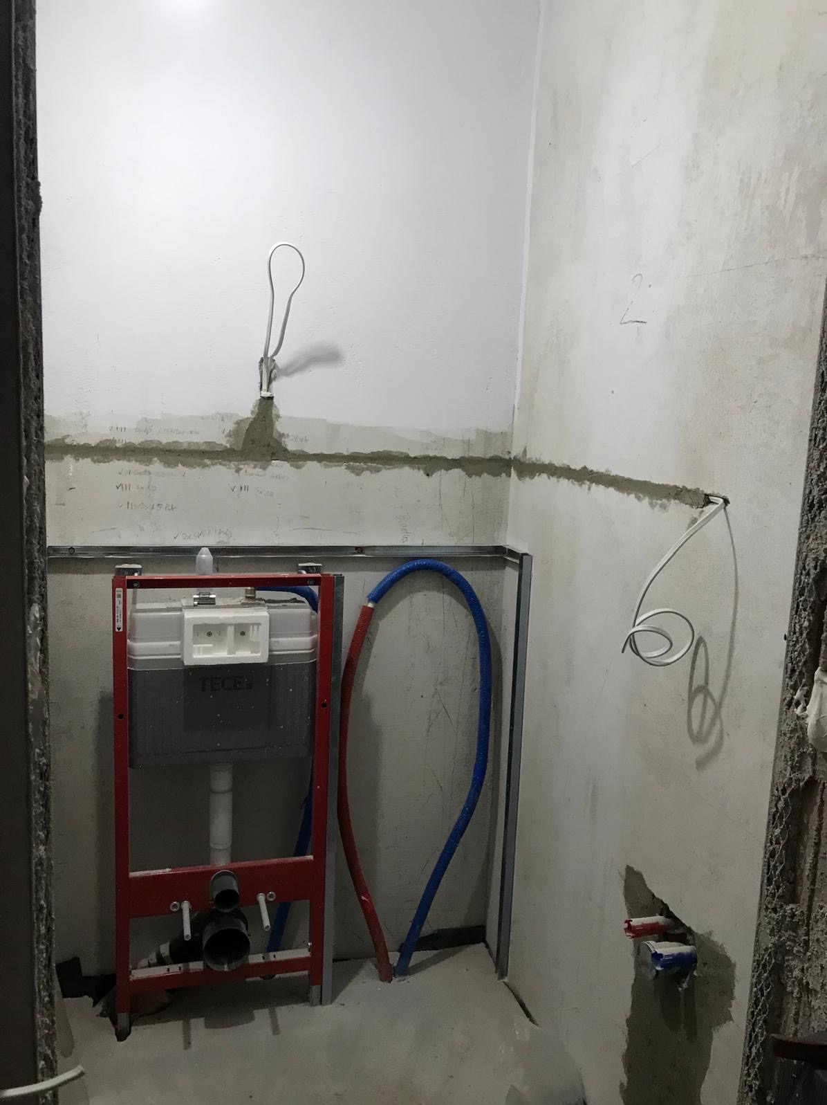 Stelaż podtynkowy WC w trakcie montażu, widoczne rury doprowadzające wodę i wyprowadzony przewód elektryczny w surowym wnętrzu łazienki.