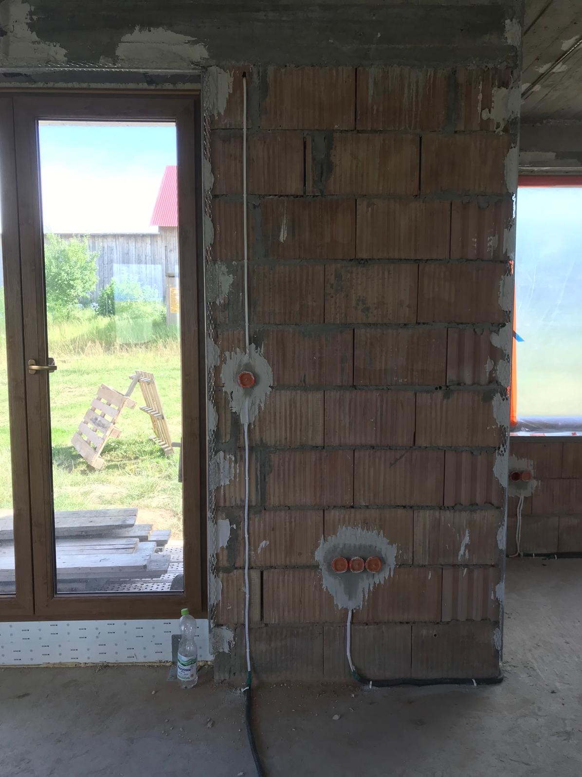 Surowa ściana z cegły z zamontowanymi puszkami elektrycznymi i przewodami. Widoczne okno z widokiem na zewnątrz. Etap prac instalacyjnych w nowym budynku.