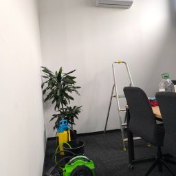 BessCool Klimatyzacja - Biała klimatyzacja AUX na ścianie biura. Obok drabina, kwiaty i sprzęt czyszczący. Usługi montażu i serwisu w Strykowie.