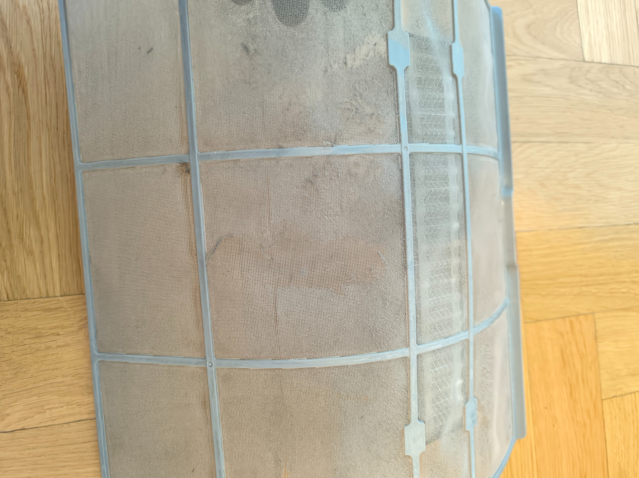 Brudny, wyjęty filtr klimatyzacji na drewnianej podłodze. Widoczna siatka i plastikowa ramka filtra, na której osiadł kurz i zabrudzenia. Perspektywa z góry.